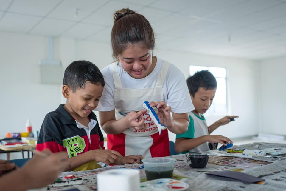 young female volunteer in red and white apron doing arts and crafts with two boys