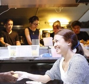woman in grey outerwear smiling with four volunteers preparing food in the background