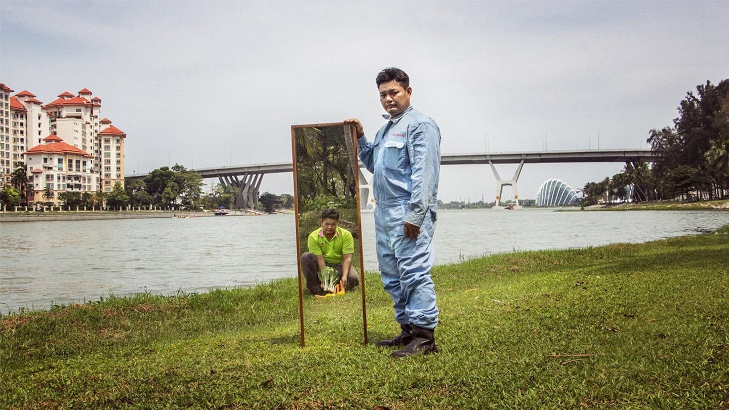 man in blue working garb holding up a mirror reflecting himself as a rubbish collector
