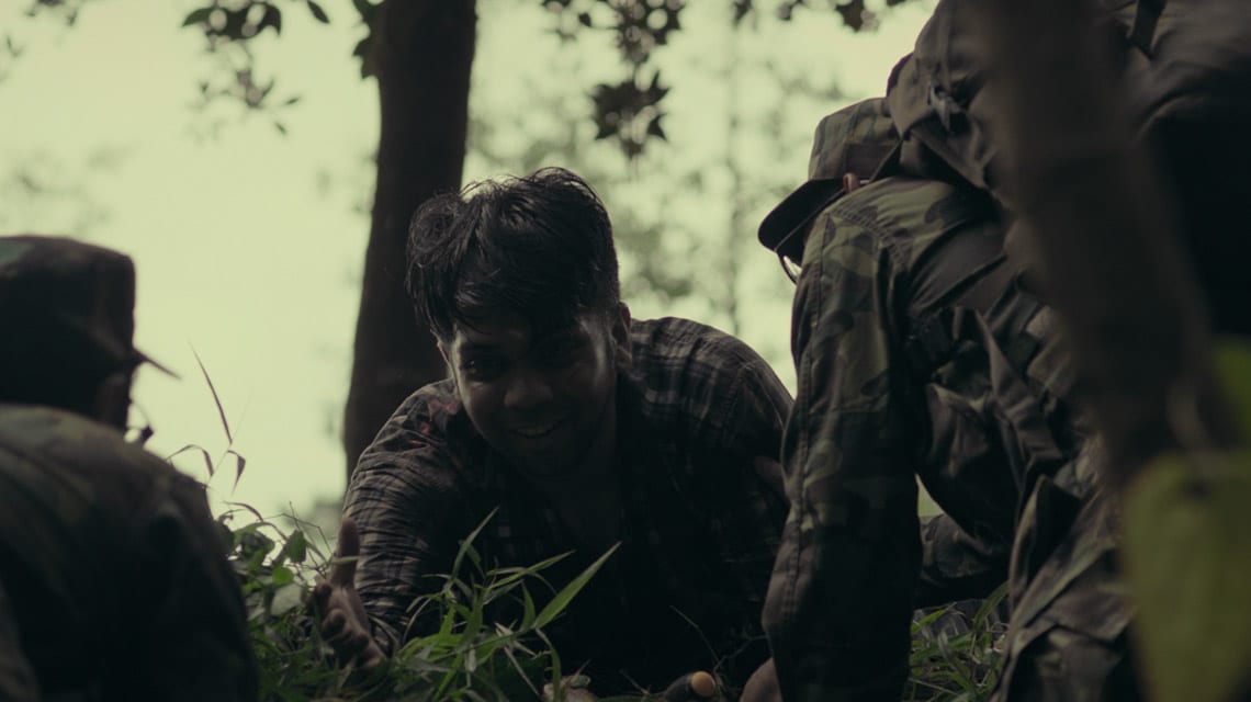 indian man in plaid shirt and two soldiers crawling through grass