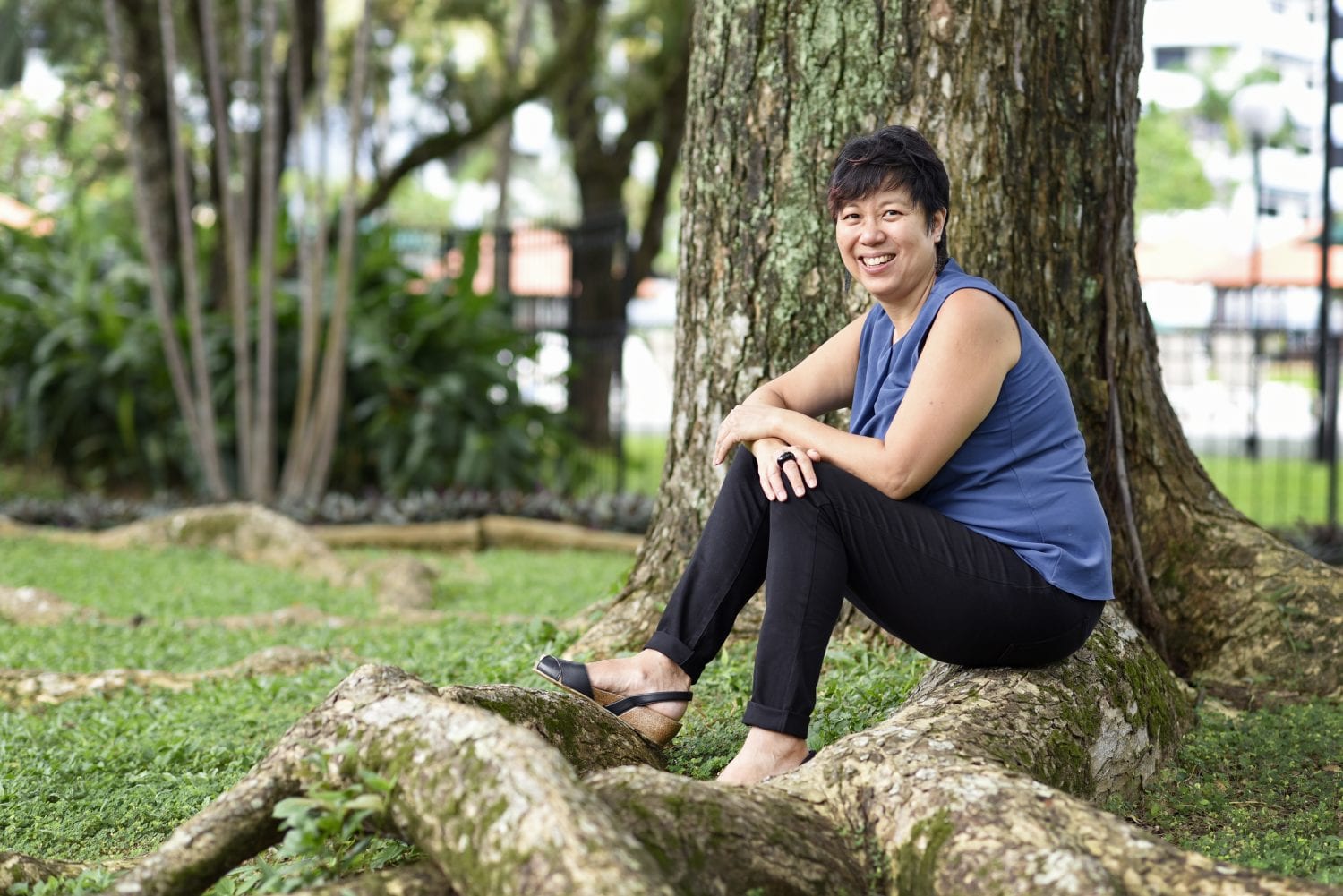 woman with pixie cut in blue sleeveless blouse and long black pants posing on a tree root
