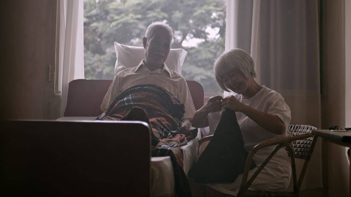 elderly man in bed and elderly woman sitting by his bed sewing