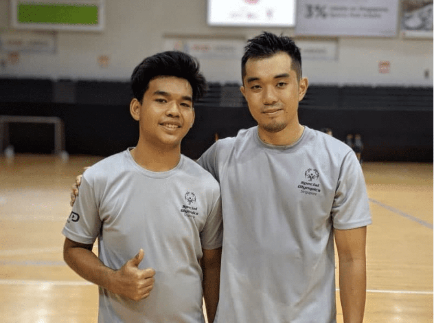 two males in grey shirts posing for the camera in an indoor sports hall