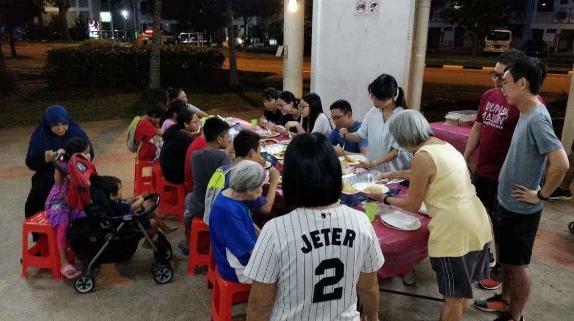 families and children having a meal around a long table while seated on red plastic chairs