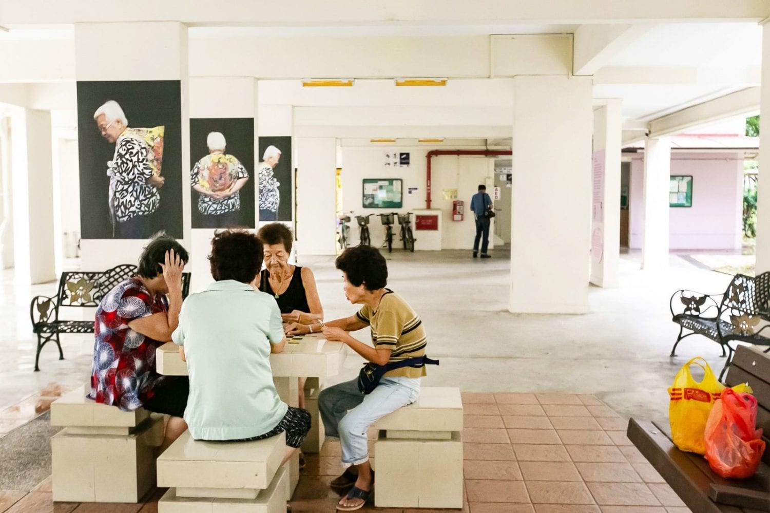 4 elderly women sitting around a void deck table with red and yellow plastic bags on bench