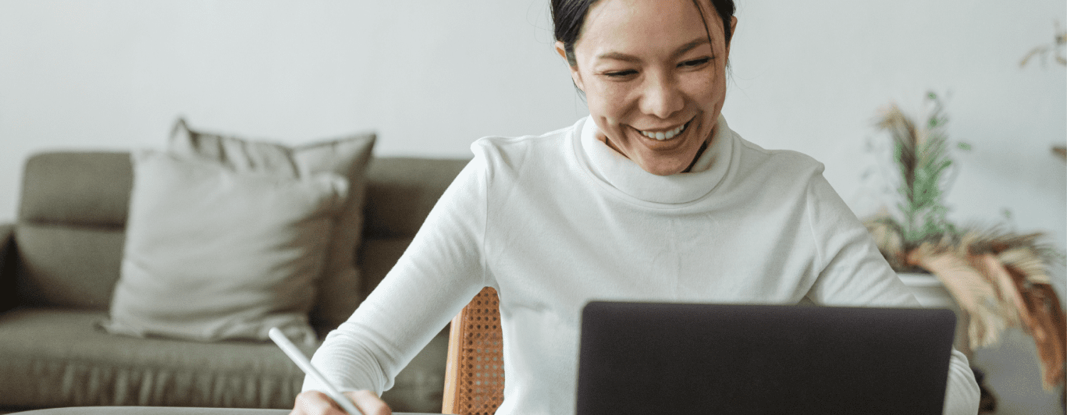 woman in long-sleeved white shirt smiling at her laptop and taking down notes