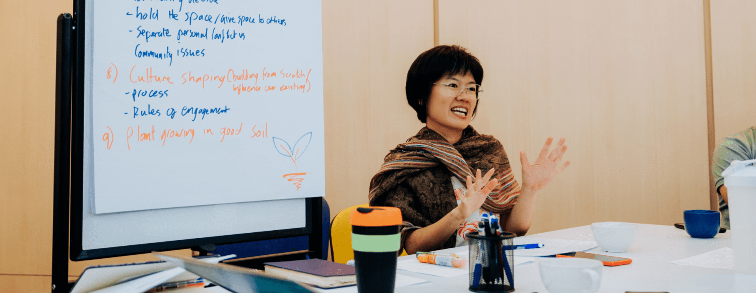 short-haired woman in brown shawl talking at a conference table in front of a whiteboard with butcher paper